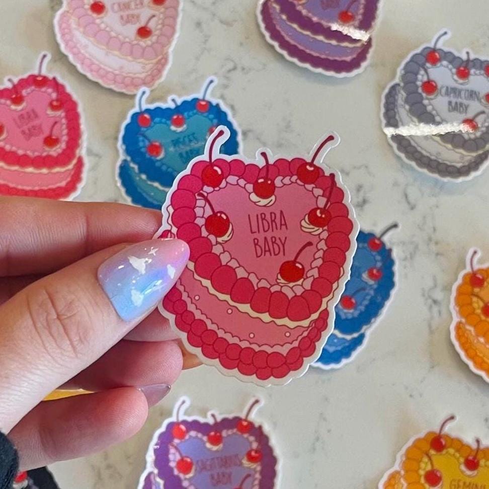 Hand holding a colorful 'Libra Baby' sticker with heart design and cherries on a marble surface.