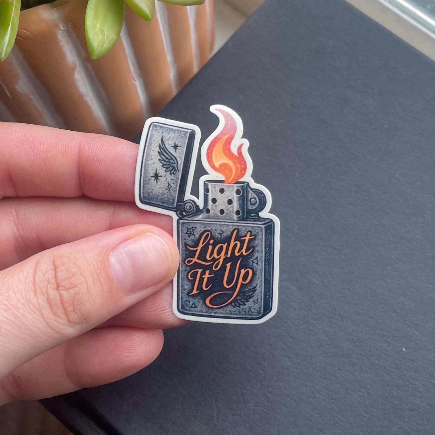 Sticker of a lighter with 'Light It Up' text held by a hand on a dark surface.