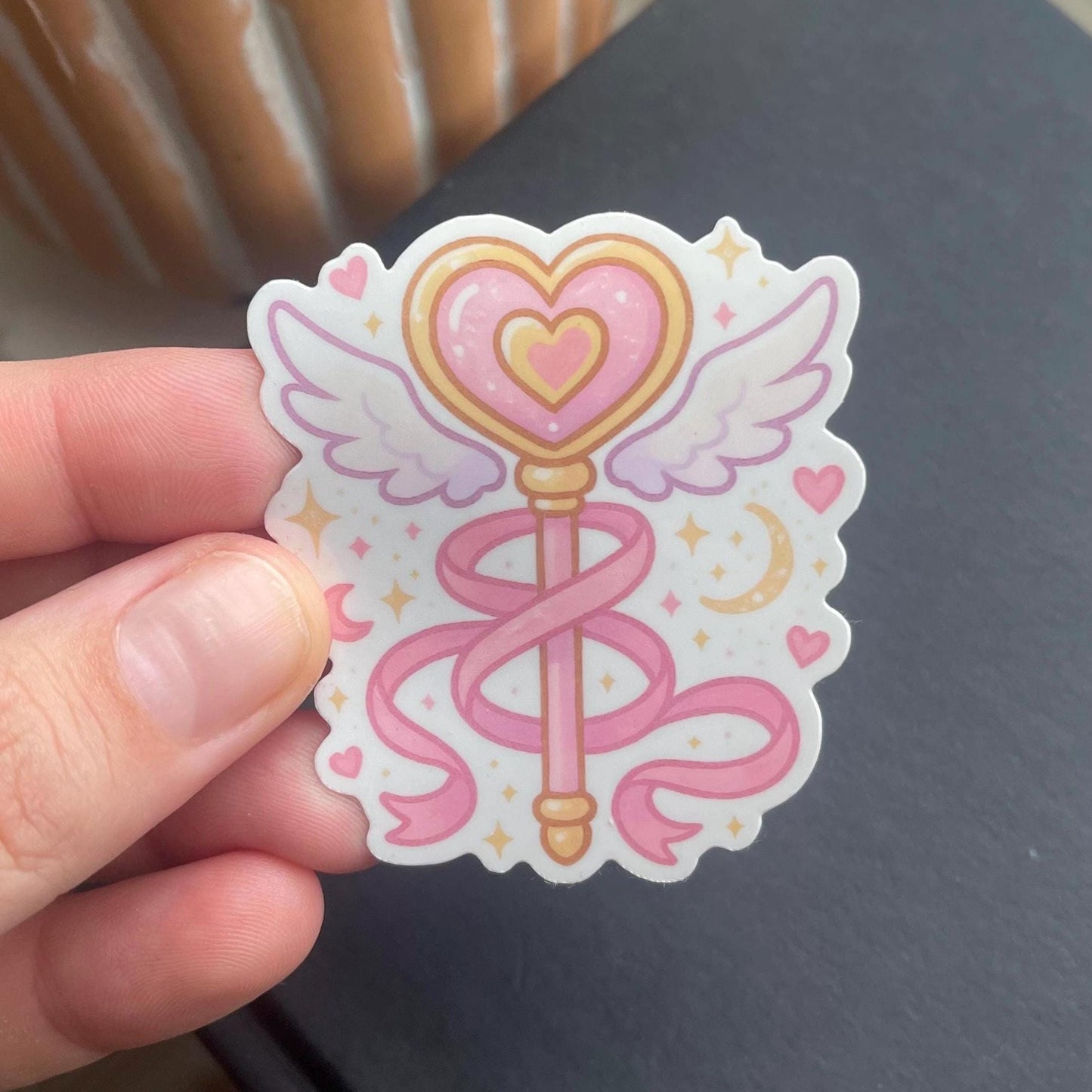 Sticker with a heart and wings wand design held by a hand on a dark surface.