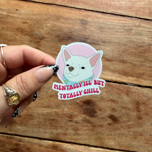 A hand with rings holds a die-cut sticker featuring a cartoon French Bulldog with the text 'MENTALLY ILL BUT TOTALLY CHILL' on a wooden surface.