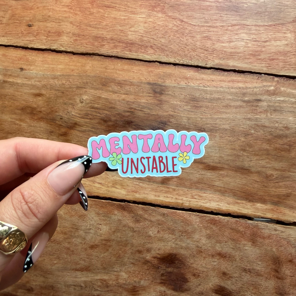 A hand with black and white patterned nail art holds a die-cut sticker that reads 'MENTALLY UNSTABLE' in pink and blue bubble letters with small yellow flowers, set against a rustic wooden background.