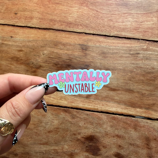 A hand with black and white patterned nail art holds a die-cut sticker that reads 'MENTALLY UNSTABLE' in pink and blue bubble letters with small yellow flowers, set against a rustic wooden background.