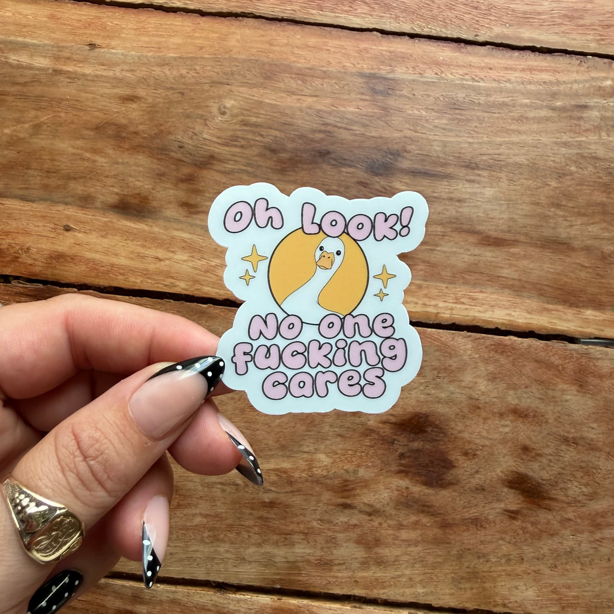 A hand with black and white patterned nails holds a die-cut sticker featuring a cartoon duck and the text 'Oh LOOK! No-one fucking cares' in pink lettering, set against a rustic wooden background.
