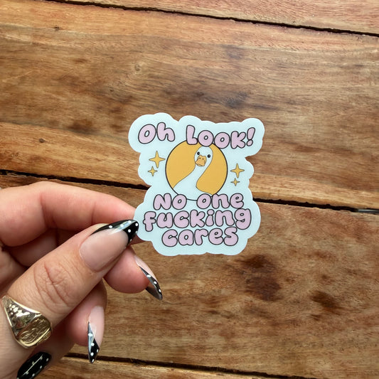 A hand with black and white patterned nails holds a die-cut sticker featuring a cartoon duck and the text 'Oh LOOK! No-one fucking cares' in pink lettering, set against a rustic wooden background.
