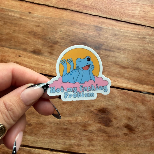 A hand with black-and-white patterned nail art holds a die-cut sticker featuring a blue cartoon frog lying on pink clouds against a yellow sun, with the text 'Not my fucking Problem' on a rustic wooden background.