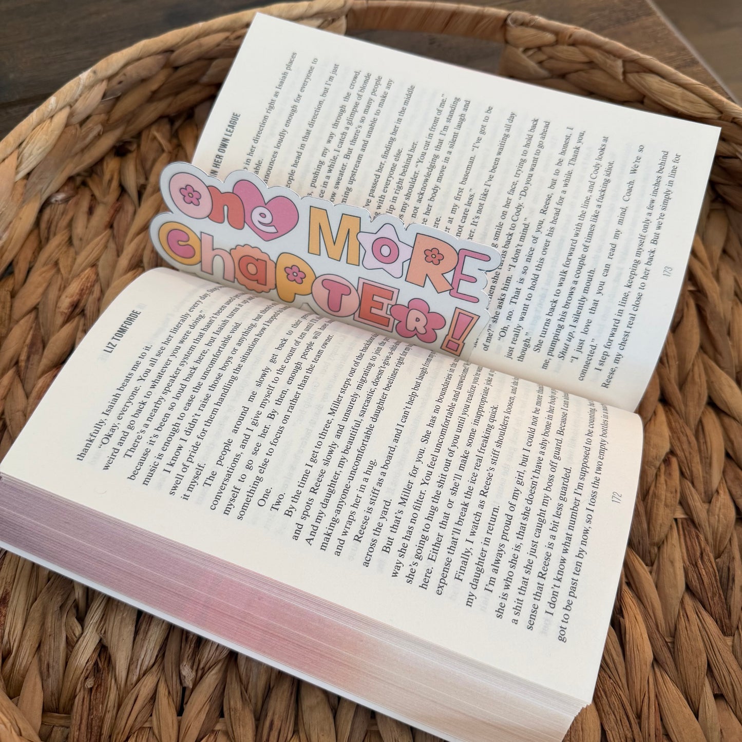 Open book with a colorful bookmark titled 'One More Chapter!' on a woven surface.