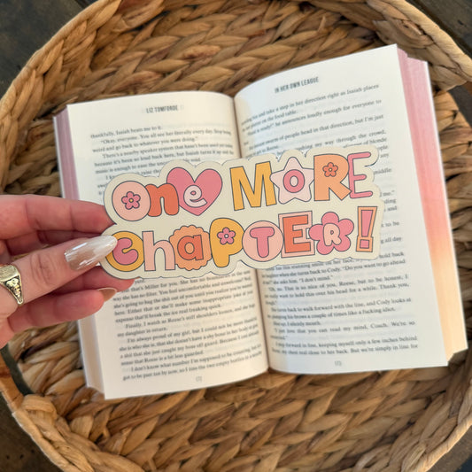 Open book with 'one more chapter!' bookmark held by a hand on a woven surface