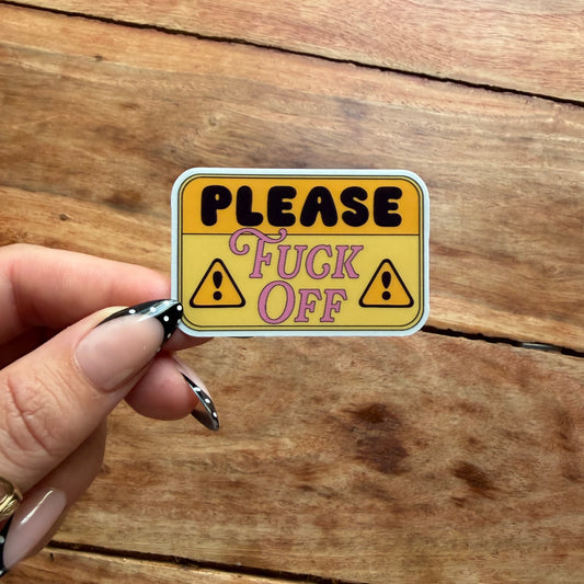 A hand with black and white patterned nail polish holds a rectangular yellow warning-style sticker that reads “PLEASE Fuck Off” with two exclamation marks, against a rustic wooden plank background.