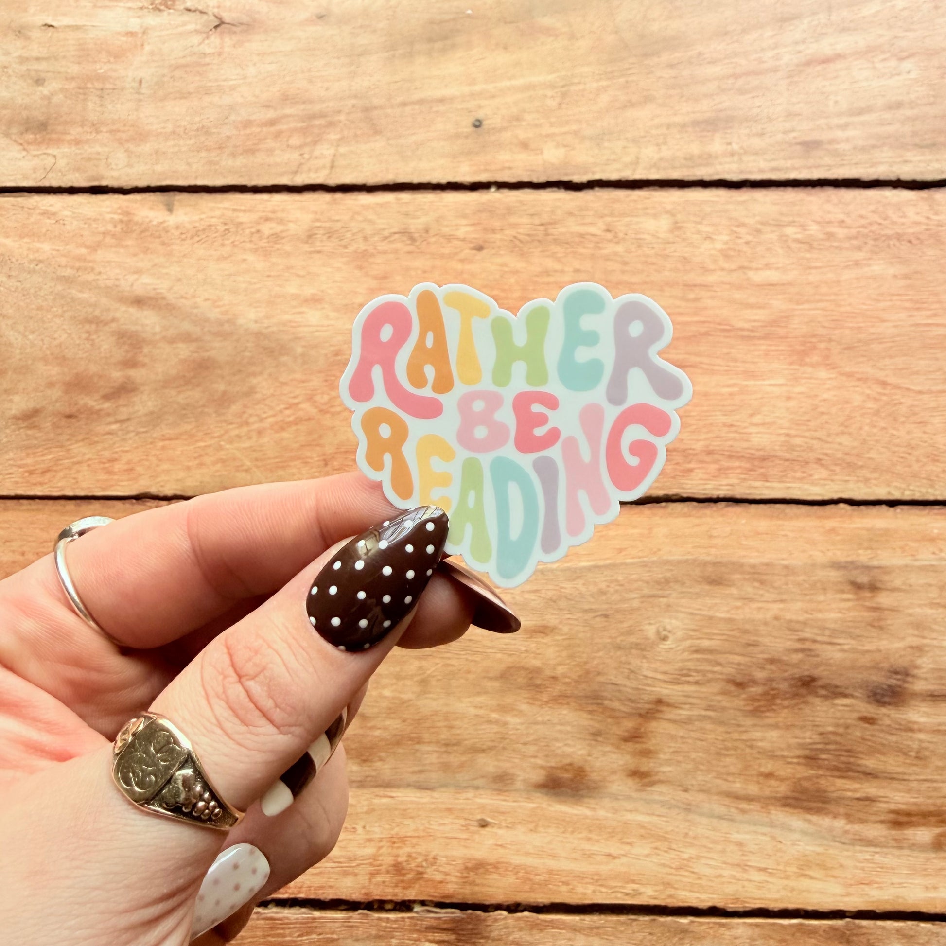 Hand holding a 'Rather Be Reading' sticker on a wooden surface