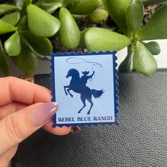 Hand holding a blue card with a horse design and 'Rebel Blue Ranch' text, against a green plant background.