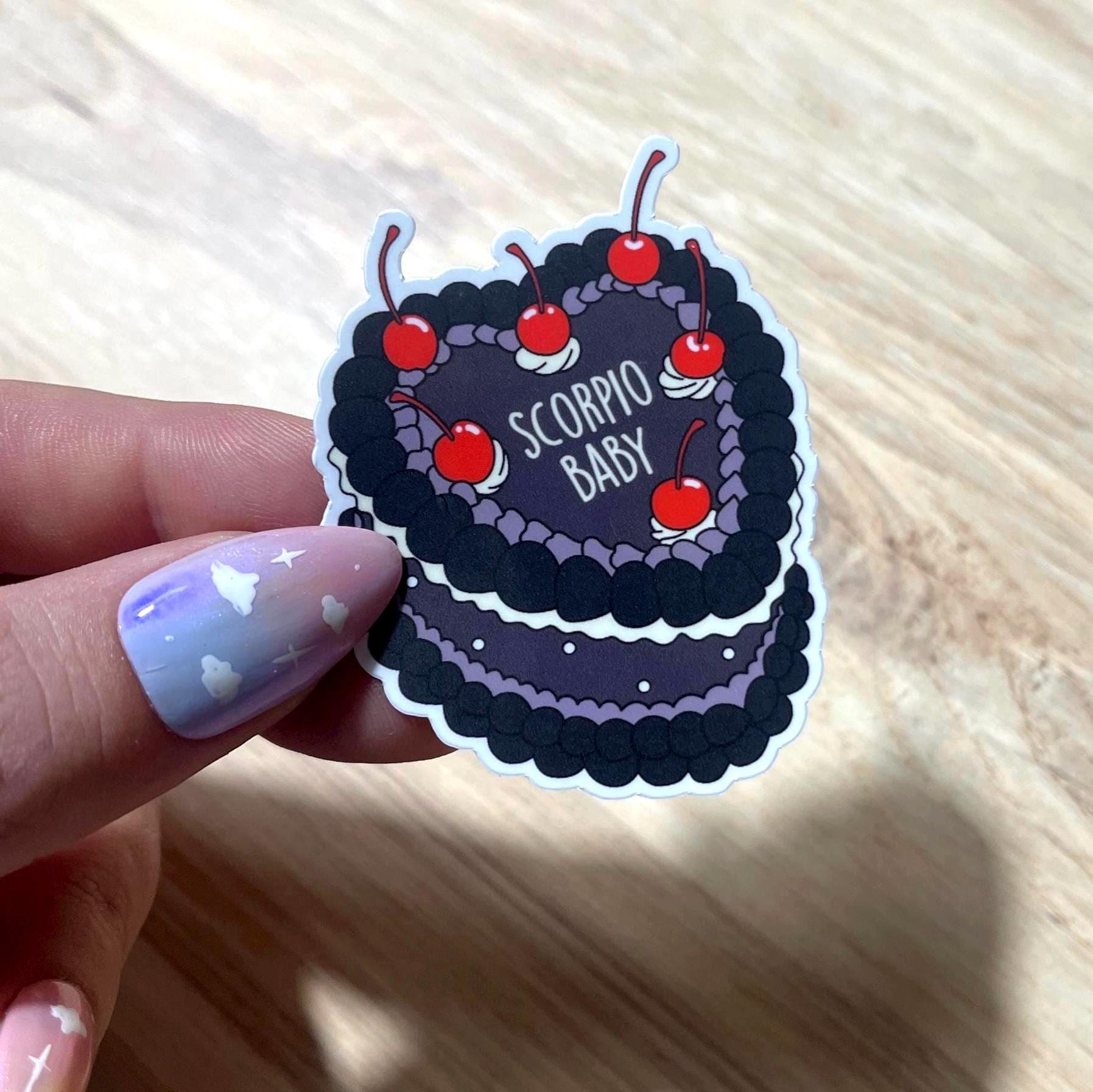 Sticker of a cake with cherries and 'Scorpio Baby' text held by a hand on a wooden surface.