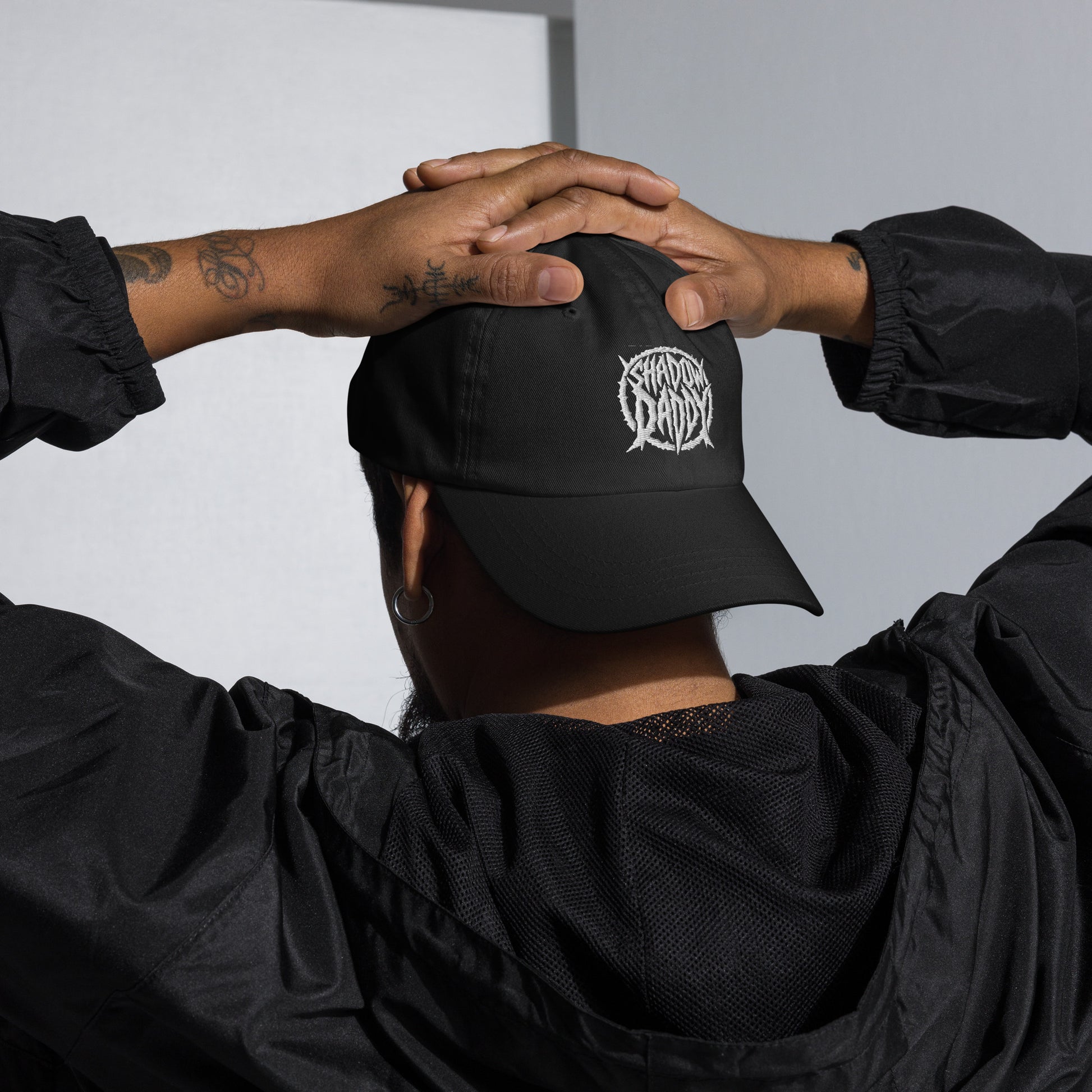 Person wearing a black cap with 'shadow daddy' logo, hands on head, against a neutral background