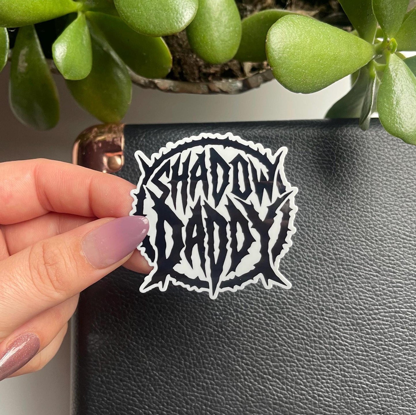 Hand holding a 'Shadow Daddy' sticker in front of a black surface with green plants.