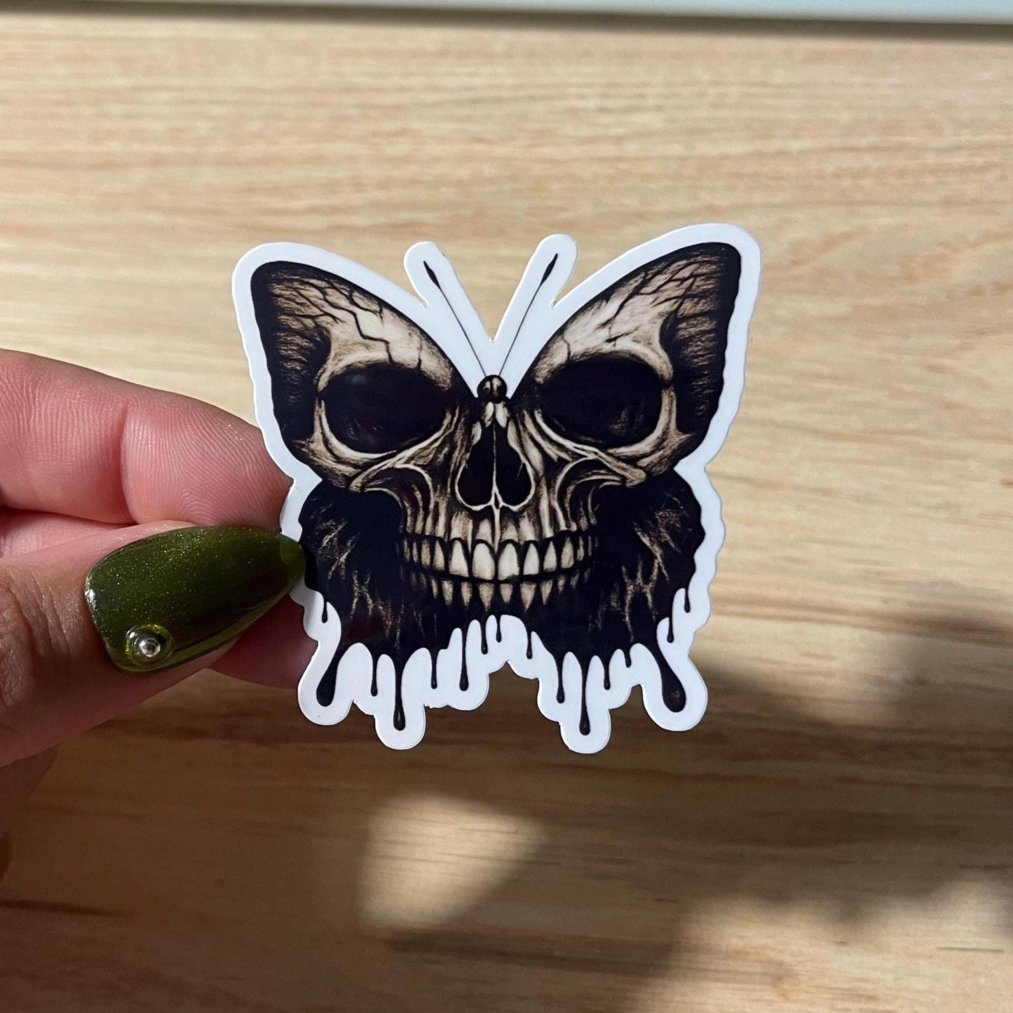 Butterfly-shaped sticker with skull design held by a hand on a wooden surface
