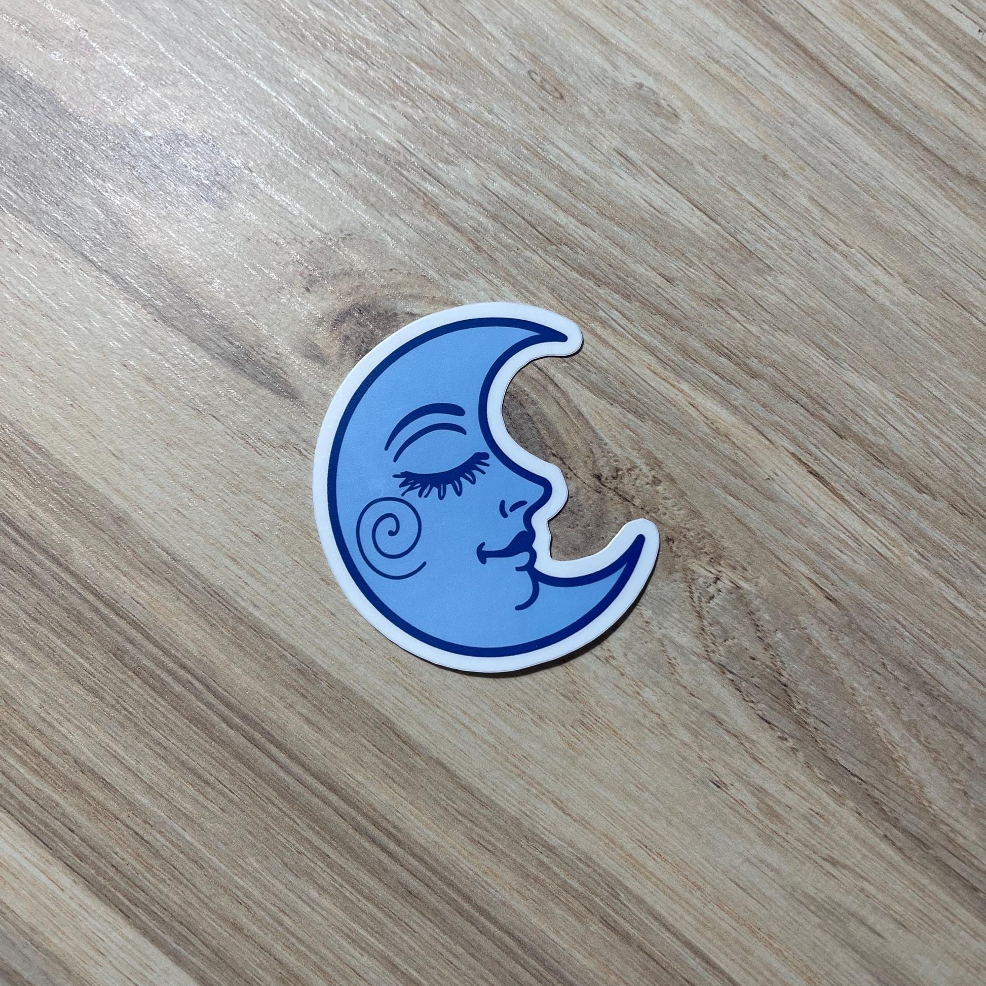 Blue moon-shaped sticker with a face design on a wooden surface