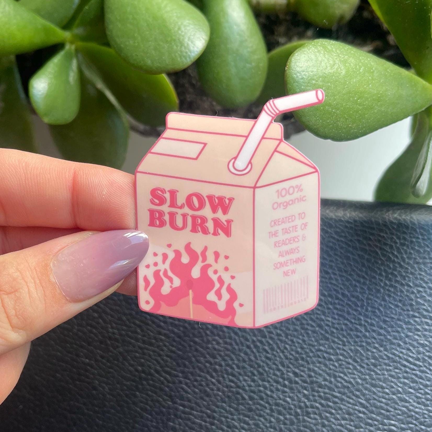 Pink 'Slow Burn' milk carton-shaped sticker held by a hand with green plants in the background.