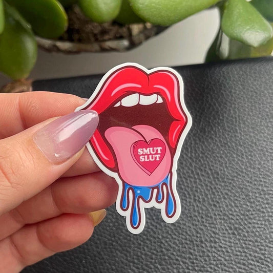 Sticker with red lips and 'Smut Slut' text held by a hand on a gray surface.