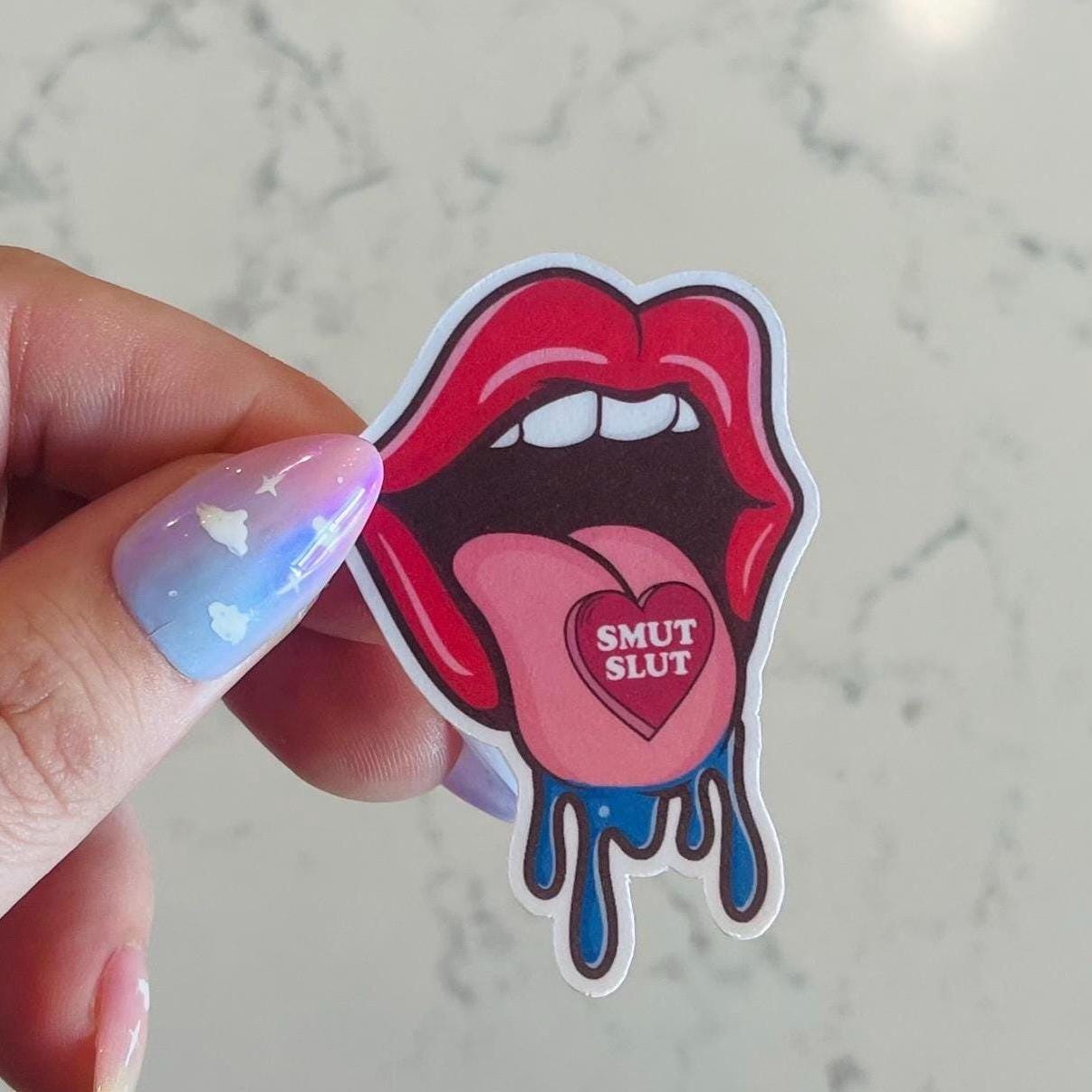Sticker of a mouth with a heart that says 'Smut Slut' held by a hand on a marble background