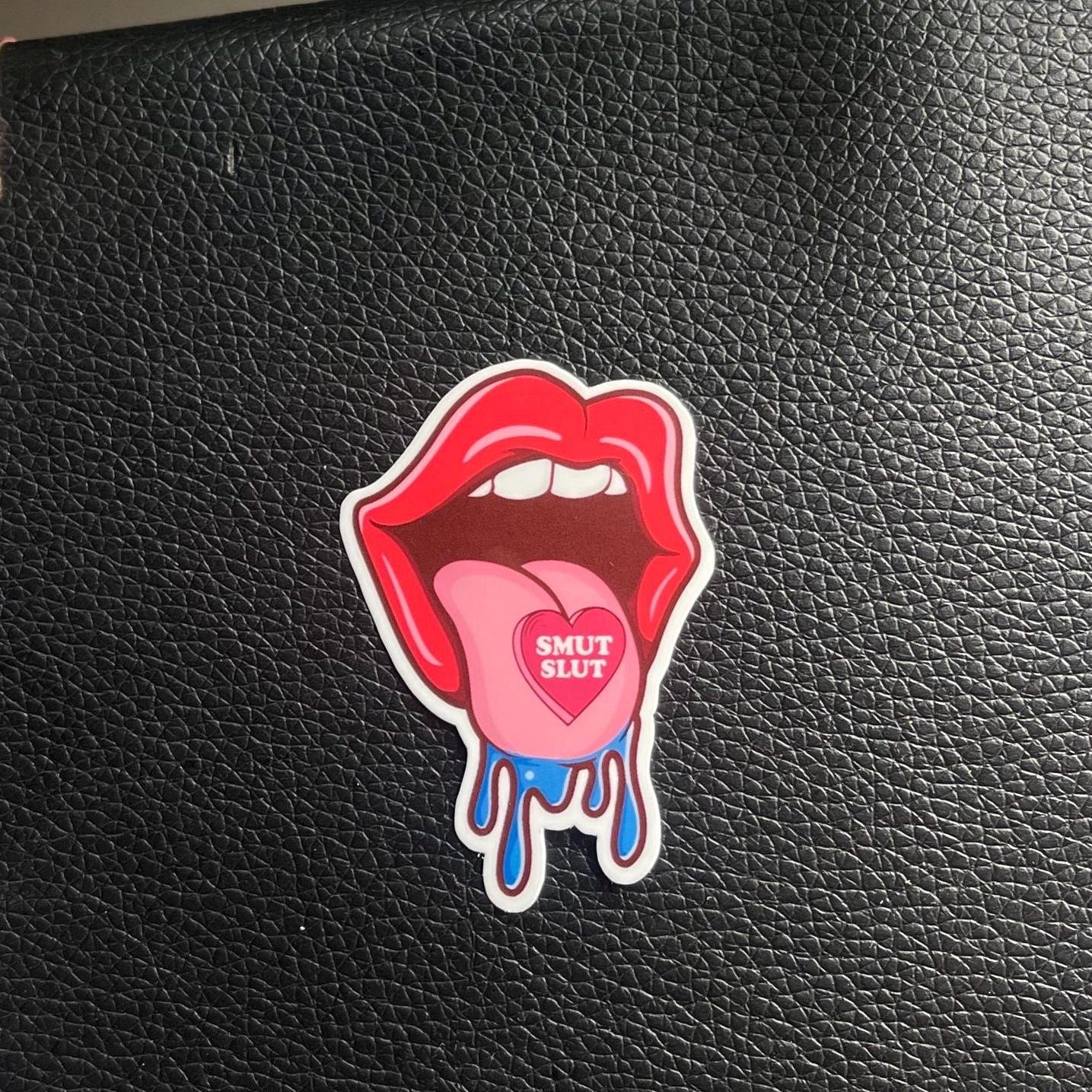 Sticker of a mouth with a tongue and 'SMUT SLUT' text on a black leather surface.
