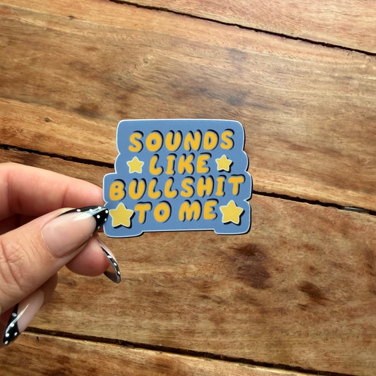 A hand with patterned nail art holds a die-cut sticker that reads 'SOUNDS LIKE BULLSHIT TO ME' in yellow letters on a blue background with yellow stars, set against a rustic wooden surface.