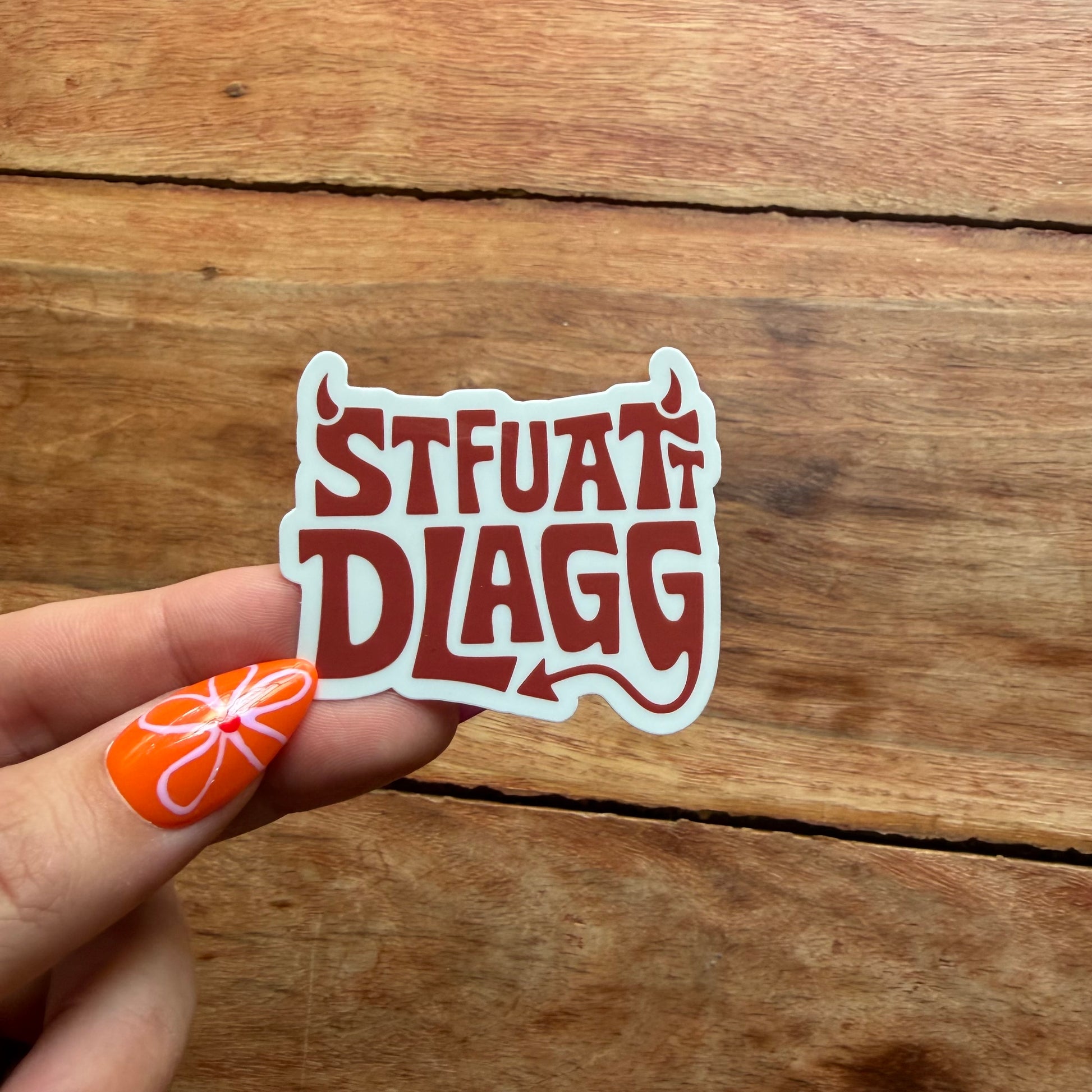 Hand holding a sticker with 'STFUAT DLAGG' text against a wooden background