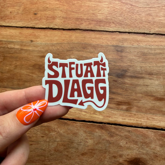 Hand holding a sticker with 'STFUAT DLAGG' text against a wooden background