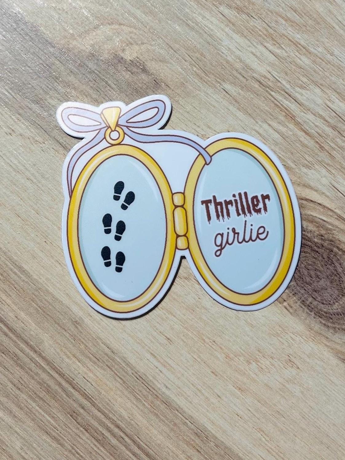 Sticker with an open locket design with "thriller girlie" on a wooden surface