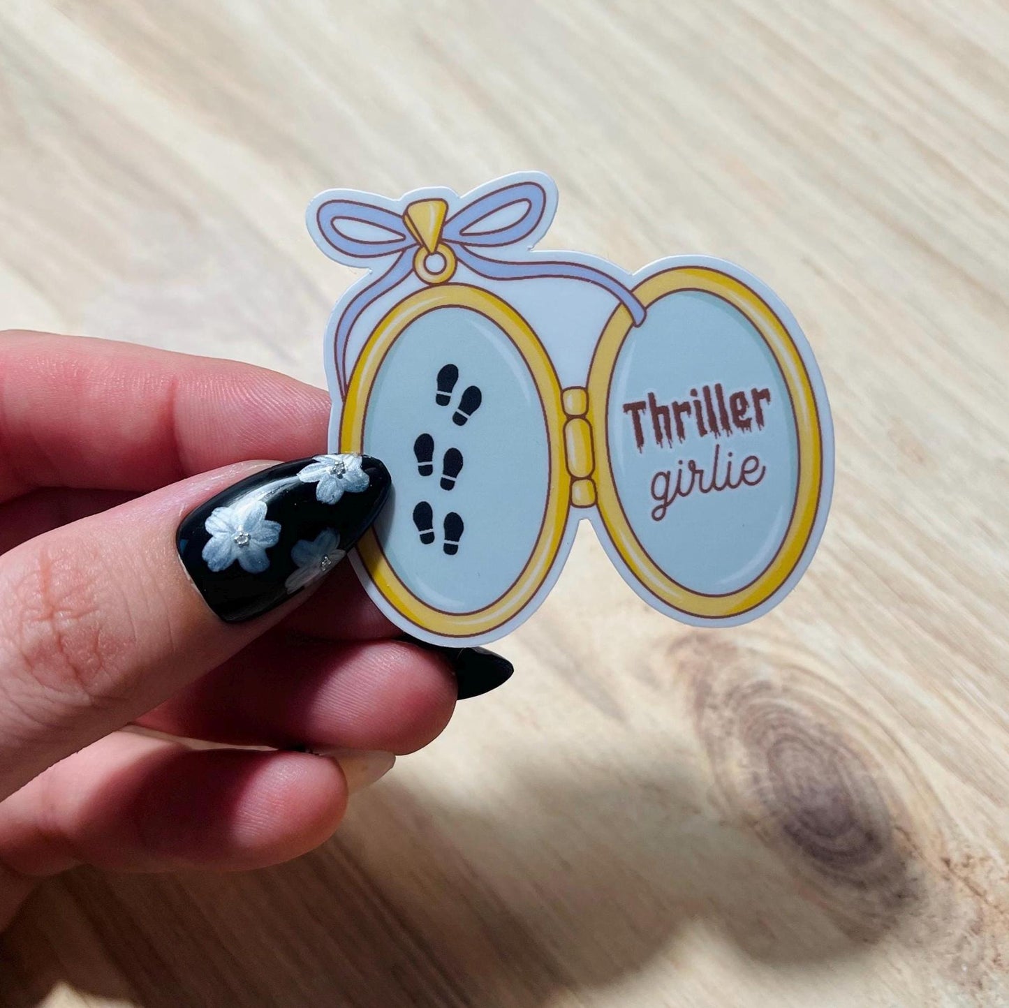 Sticker with 'Thriller girlie' text held by a hand on a wooden surface