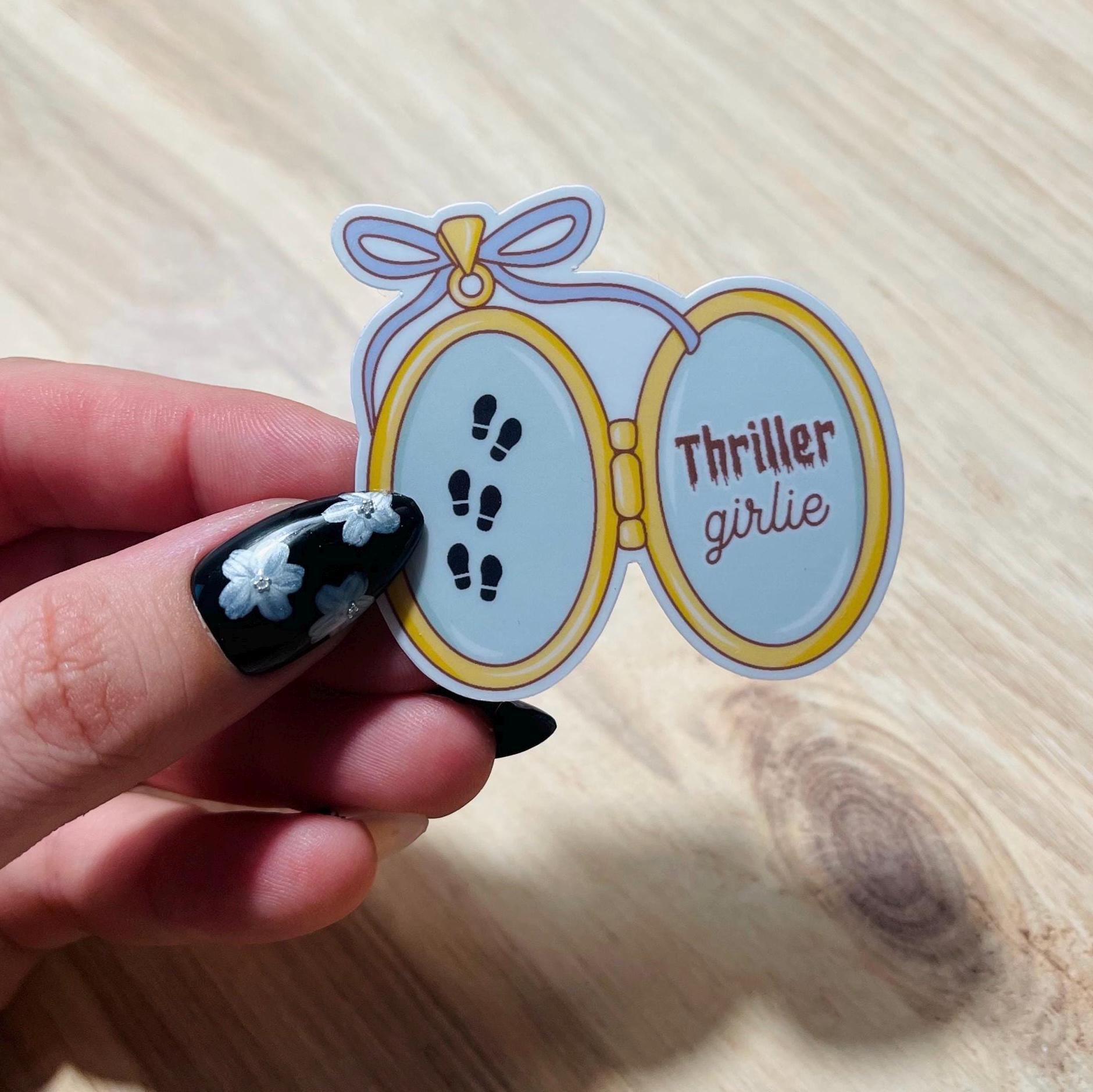 Sticker with 'Thriller girlie' text held by a hand on a wooden surface