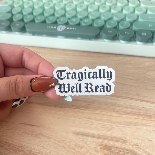 Hand holding a 'Tragically Well Read' sticker in front of a keyboard.