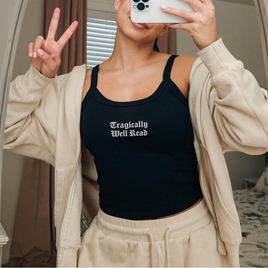 Person wearing a black tank top with 'Tragically Well Read' text, beige cardigan, and pants in front of a mirror.