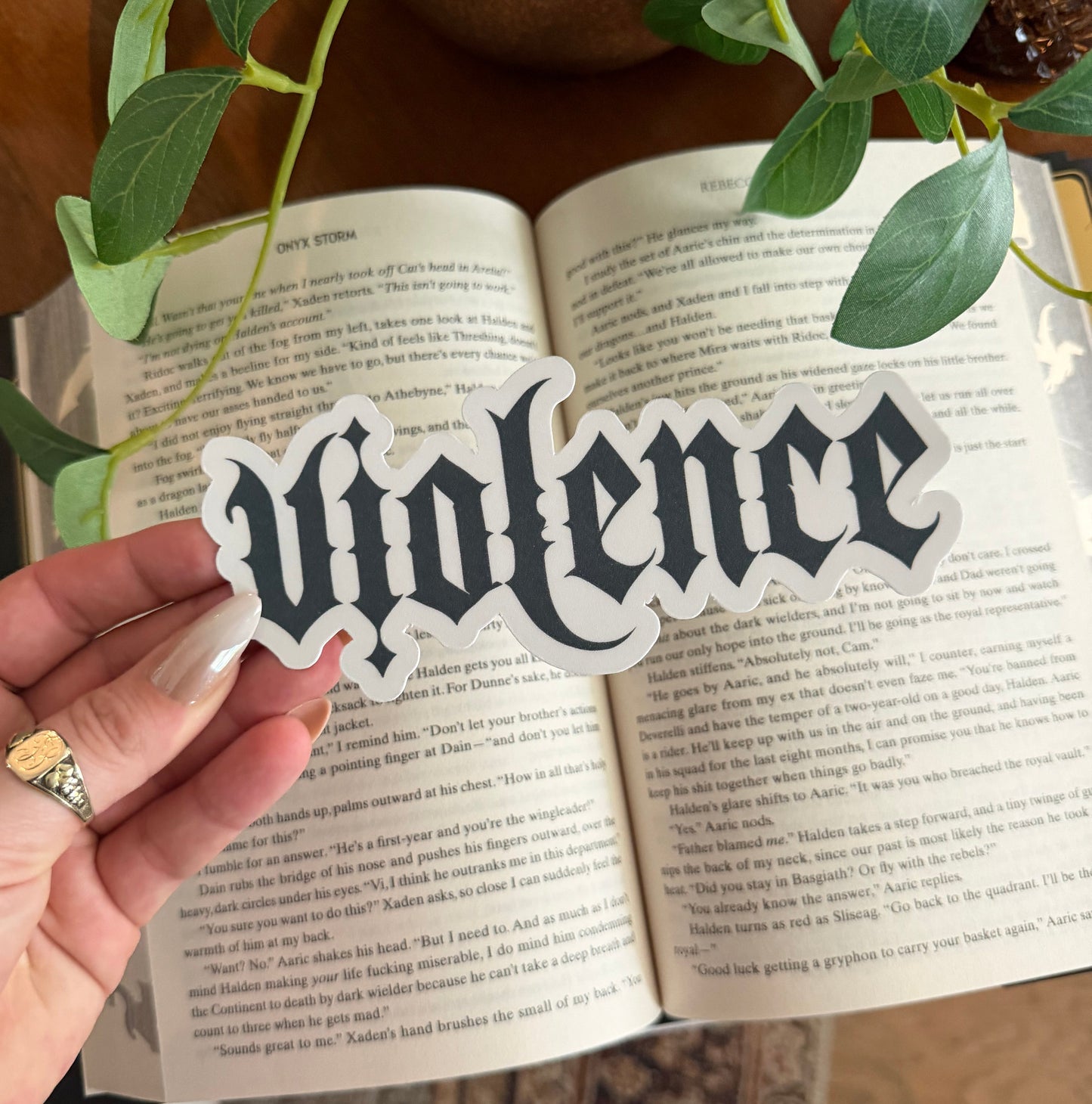 Hand holding a decorative bookmark with text over an open book, surrounded by plants.