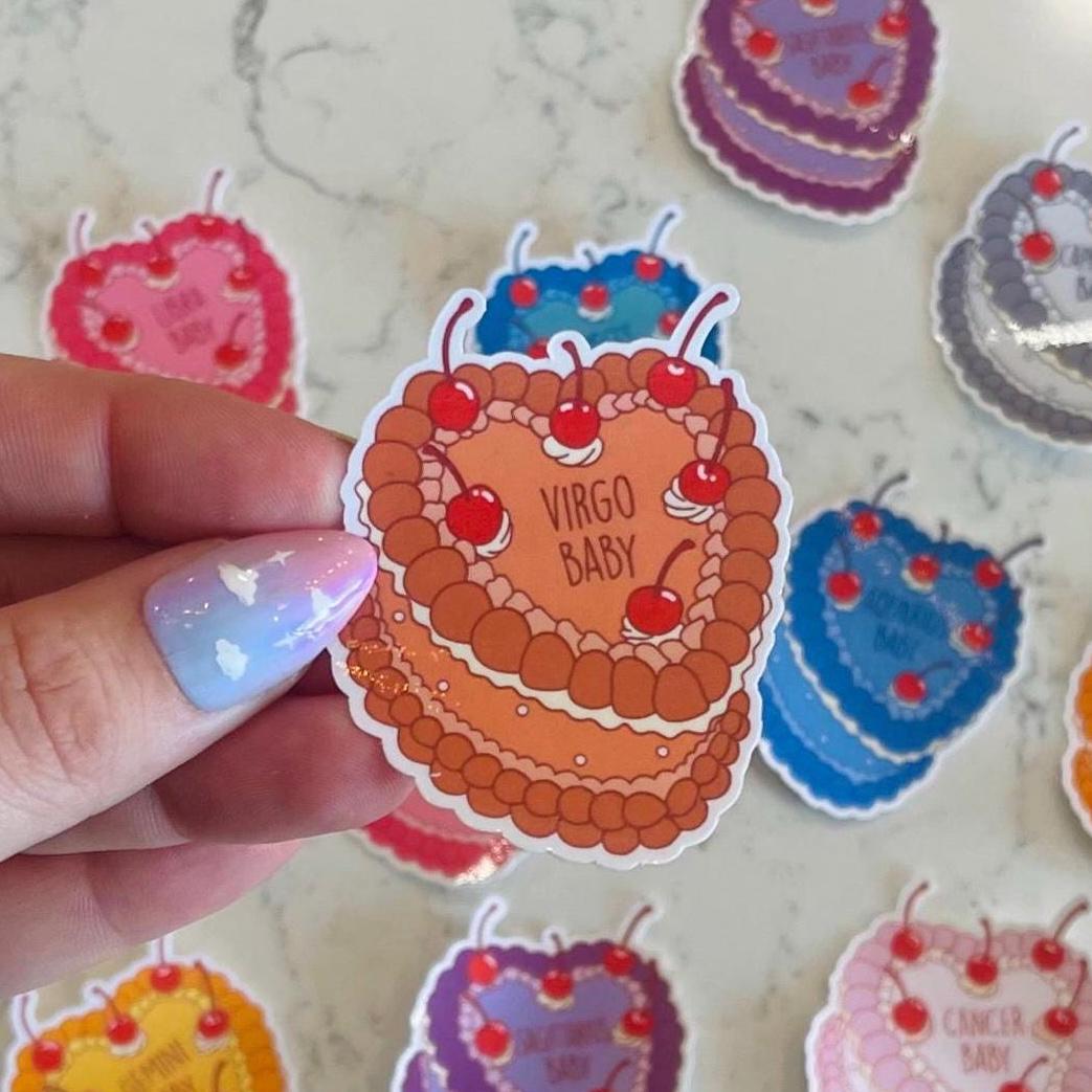 Hand holding a 'Virgo Baby' cake-shaped sticker with more similar stickers in the background.