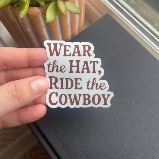 Hand holding a sticker with text 'Wear the Hat, Ride the Cowboy' against a dark surface.