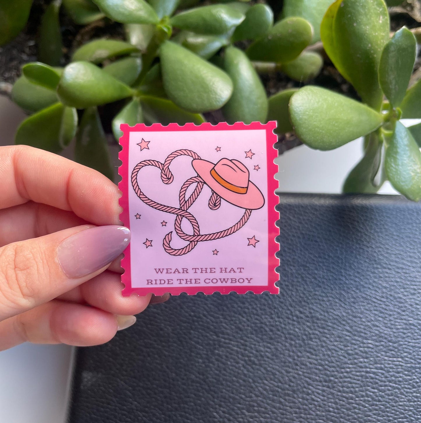Pink stamp-shaped pin with cowboy hat design held by a hand against a plant background