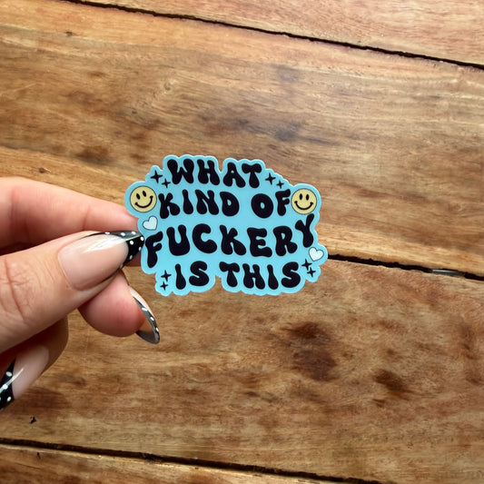 A hand with black and white patterned nail art holds a die-cut sticker with the text 'WHAT KIND OF FUCKERY IS THIS' in black lettering on a light blue background with yellow smiley faces and small star and heart accents, set against a rustic wooden surface.