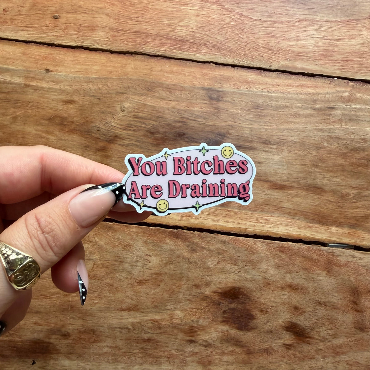 A hand with manicured nails holds a die-cut sticker that reads 'You Bitches Are Draining' in pink lettering on a light pink background with a white border, set against a rustic wooden plank surface.