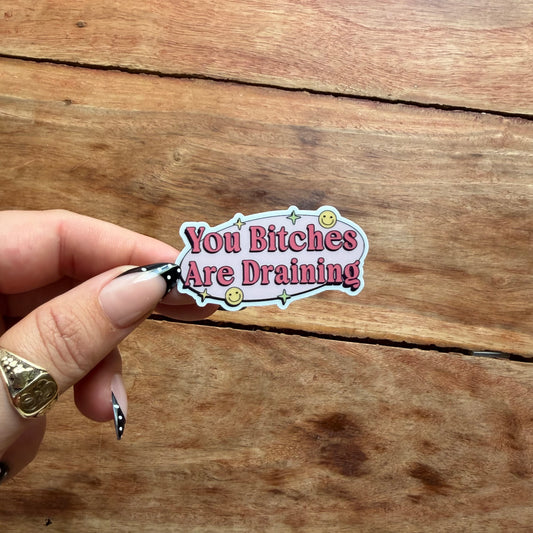 A hand with manicured nails holds a die-cut sticker that reads 'You Bitches Are Draining' in pink lettering on a light pink background with a white border, set against a rustic wooden plank surface.