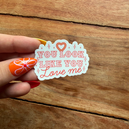a hand  holding a white sticker that says you look like you love me over a wooden surface